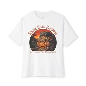 Puede incluir: Una camiseta blanca de manga corta con un gráfico de un águila negra posada sobre una calabaza tallada. El texto "EAGLE EATS PUMPKIN" aparece arriba y "CAN'T FIND MEAT? EAT WITHOUT MEAT!!!" abajo en una tipografía de color naranja y marrón.