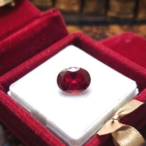 May include: A faceted oval-shaped deep red gemstone resting on a white square insert inside a red velvet jewelry box. The box features a small gold-colored ribbon detail on the corner, providing a contrast to the vibrant red velvet lining.