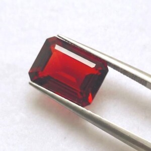 Pode incluir: Uma gema de cor vermelha profunda e vibrante com um corte esmeralda retangular, apresentando facetas nítidas e um acabamento brilhante. A pedra é segurada por uma pinça de metal prateado, exibindo sua clareza transparente e cor saturada.
