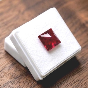May include: A square-cut, deep red gemstone with a faceted surface that reflects light. The gemstone is securely positioned in the center of a small, white, square display box with a textured white insert, showcasing its vibrant color and clarity.