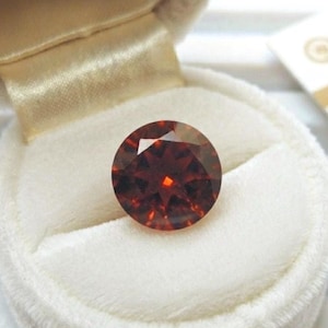 May include: A round, faceted gemstone with a deep reddish-brown color, featuring a brilliant cut with multiple triangular facets that reflect light. The gemstone is nestled securely within a plush, cream-colored velvet ring box.