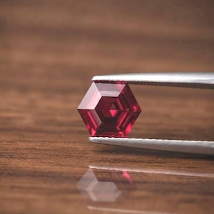 Puede incluir: Una gema de color rojo intenso y vibrante tallada en una forma hexagonal precisa. La piedra presenta múltiples facetas internas que crean un patrón geométrico, sostenida por unas pinzas de metal plateado con un acabado liso y pulido.