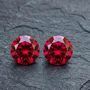 Puede incluir: Dos piedras preciosas redondas de talla brillante con un color rojo intenso y vibrante. Cada piedra presenta una superficie compleja de múltiples facetas que refleja la luz, creando un patrón simétrico de facetas triangulares.