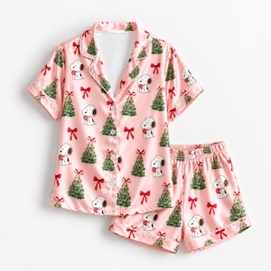May include: A light pink pajama set consisting of a short-sleeved button-down shirt and matching shorts. The fabric features a repeating pattern of Snoopy characters, green Christmas trees, and red bows with white piping along the edges.