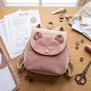 Op de afbeelding: Een kleine rugzak van beige en lichtroze corduroy met een berenvormige flap, twee afgeronde oren en een houten knoopsluiting. De rugzak ligt tussen naaipatronen met de tekst Mini-Bär, stof, knopen en een schaar.
