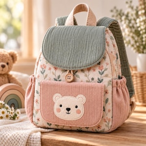Könnte beinhalten: Ein kleiner Rucksack mit Blumenmuster auf cremefarbenem Grund, einem salbeigrünen Deckel und einer rosafarbenen Vordertasche. Die Vordertasche zeigt ein aufgesticktes Bärengesicht. Der Rucksack hat einen Tragegriff, verstellbare Schultergurte und einen Knopf.