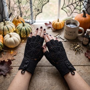 May include: A pair of black fingerless gloves featuring a crocheted spiderweb pattern on the back of the hand, a textured wrist section, and two small black buttons near the cuff. The gloves are crafted with a loop detail at the wrist edge.