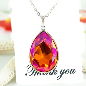 Può includere: Una collana con catena d'argento con un ciondolo a forma di lacrima con un grande cristallo sfaccettato arancione e rosa. Il ciondolo è su una carta bianca con il testo "Grazie".