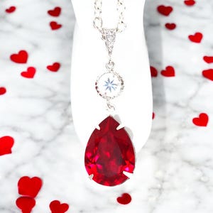 May include: A silver chain necklace with a teardrop-shaped red crystal pendant and a smaller clear crystal pendant. The necklace is on a white background with red hearts scattered around it.
