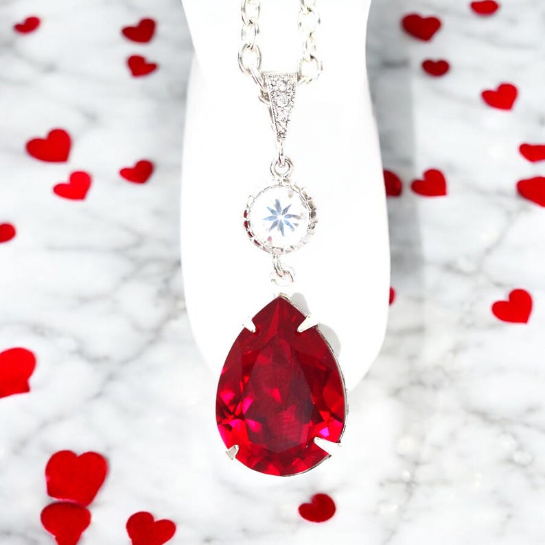 May include: A silver chain necklace with a teardrop-shaped red crystal pendant and a smaller clear crystal pendant. The necklace is on a white background with red hearts scattered around it.