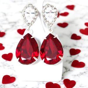 May include: A pair of silver teardrop earrings with a large red crystal and a smaller clear crystal halo.