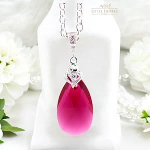 May include: A silver-toned necklace with a large, teardrop-shaped, fuchsia-colored pendant. The pendant is suspended from a delicate chain and features a decorative, crystal-studded bail. The necklace is displayed against a white background.