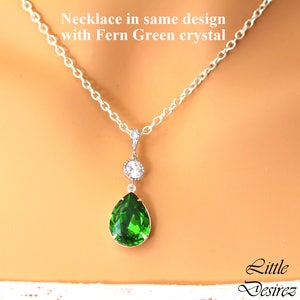 May include: A silver chain necklace with a teardrop-shaped pendant featuring a large, bright green crystal and a smaller clear crystal above it. The text "Necklace in same design with Fern Green crystal" is visible above the necklace.