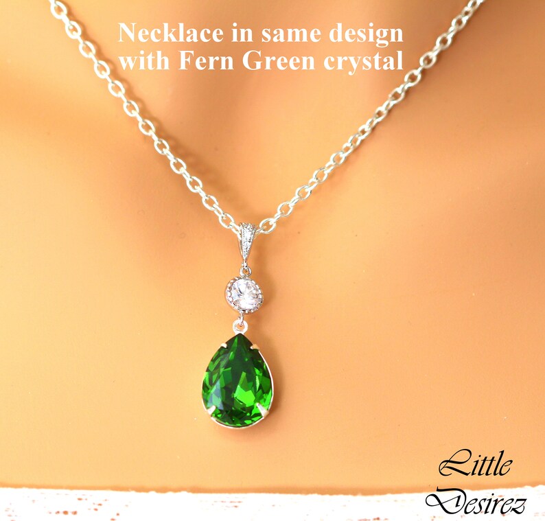 May include: A silver chain necklace with a teardrop-shaped pendant featuring a large, bright green crystal and a smaller clear crystal above it. The text "Necklace in same design with Fern Green crystal" is visible above the necklace.