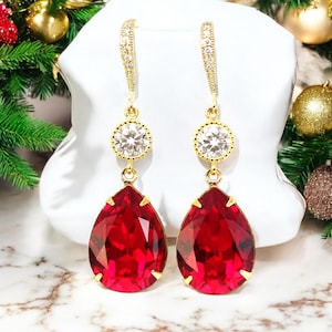 Op de afbeelding: Een paar gouden oorhangers met een grote rode traanvormige kristal en een kleinere ronde, heldere kristal. De oorbellen hebben een delicate gouden ketting met kleine heldere kristallen.