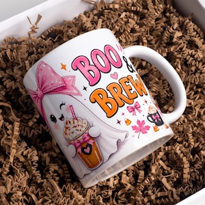 May include: A white ceramic mug featuring a cartoon ghost wearing a pink bow and holding a coffee cup. The text BOO & BREW is printed in large pink and orange letters. Additional graphics include small pink bows, maple leaves, and candy corn.