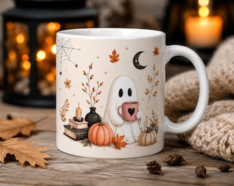 May include: A white ceramic mug featuring a printed illustration of a white ghost holding a pink mug with a black heart. The design includes orange pumpkins, a stack of books with a lit candle, autumn leaves, a spider web, and a crescent moon.