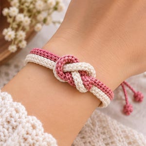 May include: A dual-coloured bracelet featuring a woven cord design in dusty rose and off-white. The centrepiece is a prominent infinity-style knot where the two coloured cords intertwine. The bracelet includes adjustable pull strings with small knotted ends.