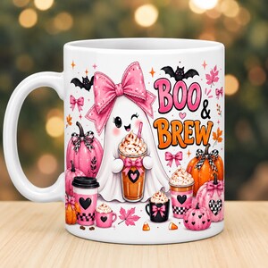 May include: A white ceramic mug featuring a cartoon ghost wearing a large pink bow and holding a coffee cup. The design includes orange and pink pumpkins, bats, maple leaves, and the text "BOO & BREW" in orange and pink lettering.