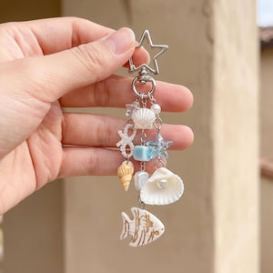 Puede incluir: Un llavero plateado en forma de estrella con una colección de dijes colgantes que incluyen conchas blancas, una estrella azul translúcida, un pequeño cubo azul, una concha en espiral, una perla blanca y un pez plano blanco con líneas marrones.