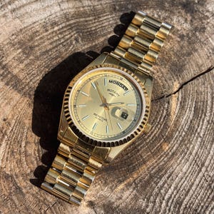 May include: A gold-tone Rotary quartz wristwatch with a fluted bezel and a matching link bracelet. The gold dial features a day display at the twelve o'clock position, a date window at the three o'clock position, and the text 100 METERS QUARTZ.