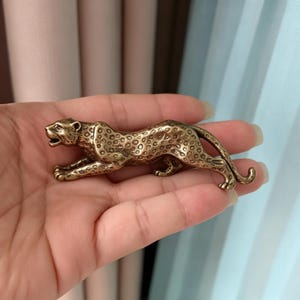 以下が含まれることがあります： 獲物を狙うヒョウの形をした、ブロンズ色の小さな金属製フィギュア。彫刻の表面には細かな質感があり、体全体にエンボス加工された斑点模様が施されています。口は開いており、長い尾が後ろに伸びています。