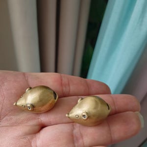 May include: Two small, metallic gold-colored mouse-shaped figurines. Each piece features a smooth, rounded body, a pointed snout, and small, defined ears. The surface has a brushed, matte finish, and the overall design is simple and compact.