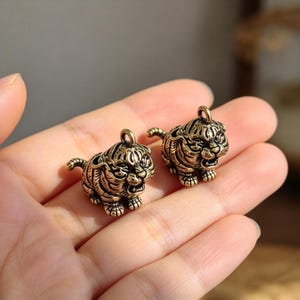 Può includere: Due piccoli ciondoli a forma di tigre in ottone in stile antico, ognuno raffigurante una tigre accovacciata con la bocca aperta, strisce visibili e un piccolo anello di metallo sulla parte superiore per l'aggancio, con finitura dorata e superficie ruvida.