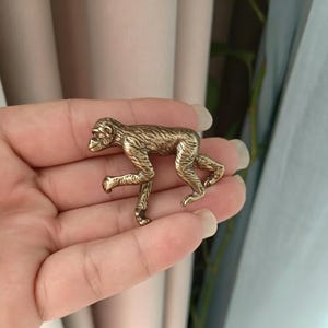 Könnte beinhalten: Eine kleine, metallene, messingfarbene Affenfigur in einer laufenden Pose. Die Oberfläche des Affen weist detaillierte, texturierte Linien auf, die Fell imitieren, während Gesicht und Gliedmaßen eine glatte Oberfläche aufweisen.