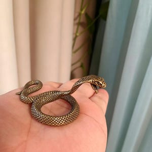 May include: A small, metallic bronze-coloured snake figurine with a textured, scale-like surface. The snake is depicted in a coiled position with its head raised, mouth slightly open, and detailed markings visible along its body and hood.