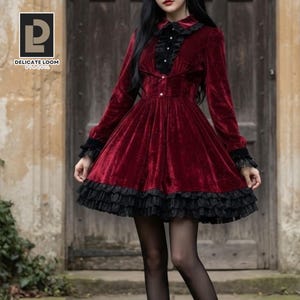 May include: A deep red velvet dress featuring long sleeves with black lace cuffs, a black lace-trimmed collar, and a button-down bodice. The skirt is flared with a layered black lace ruffle hem. The logo DELICATE LOOM BY VAQUIL is visible.