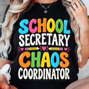 May include: A black t-shirt featuring the colorful text SCHOOL SECRETARY CHAOS COORDINATOR in a bold, rounded font. The design includes two yellow pencils and small pink hearts centered between the words, all printed in bright, vibrant colors.