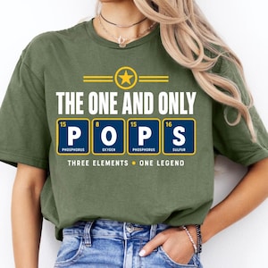 Puede incluir: Una camiseta verde oliva con un gráfico que incluye el texto THE ONE AND ONLY POPS. Debajo, cuatro cuadrados azules representan elementos químicos con las letras P, O, P, S, números atómicos y nombres. El texto THREE ELEMENTS ONE LEGEND aparece debajo.