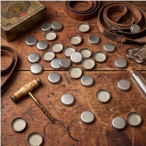 May include: A collection of silver-colored metal bottle caps scattered across a wooden surface, accompanied by a corkscrew with a light-colored handle, a small glass vial, a vintage metal tin, and coiled brown leather straps.