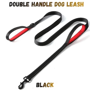 Pode incluir: Uma trela preta para cão com duas pegas, apresentando um forro interior vermelho vibrante nas alças das pegas e costuras refletoras ao longo do comprimento. A trela inclui um gancho giratório de metal preto e o texto DOUBLE HANDLE DOG LEASH BLACK.