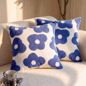 May include: Two square throw cushions featuring a white base with a repeating pattern of blue tufted flowers. Each flower has a solid blue petal shape and a white circular centre. The texture of the blue floral design is raised and plush.