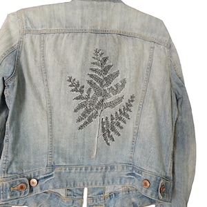 May include: A light blue denim jacket featuring a black embroidered fern leaf design on the back. The jacket has a classic collar, long sleeves, buttoned cuffs, and a buttoned waistband with metallic copper-coloured buttons on the lower hem.