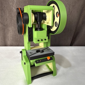 May include: A lime green mechanical punch press model featuring a large side flywheel, a black central ram assembly, and a dark gray rectangular work table. The machine includes an orange belt, black accents, and a small control panel with an orange button.