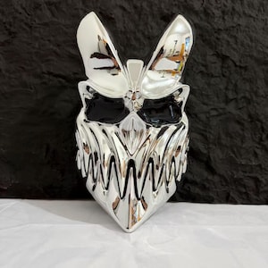 Pode incluir: Uma máscara refletora com acabamento cromado, apresentando um design de criatura estilizada e agressiva com orelhas pontiagudas, órbitas oculares escuras e profundas, e uma boca proeminente e serrilhada cheia de dentes triangulares afiados.