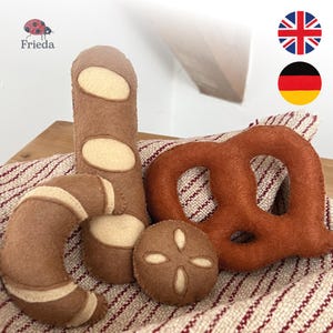 Puede incluir: Un conjunto de cuatro piezas de fieltro para jugar, que incluye una baguette marrón con cortes color crema, un panecillo en forma de media luna, un bollo redondo con un patrón en cruz y un pretzel marrón sobre una tela a rayas beige y roja.