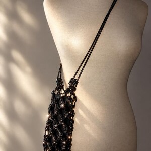 May include: A black crossbody bag featuring a decorative lattice overlay made of dark, reflective beads. The bag has a long, thin strap and a structured rectangular shape, with the beaded netting creating a shimmering, textured appearance.