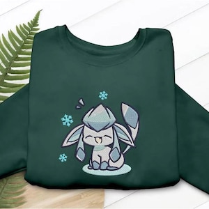May include: A dark green crewneck sweatshirt featuring an embroidered graphic of a light blue and white stylized creature with large ears and a tail, surrounded by three small light blue snowflakes, with the creature sitting on a light blue oval.