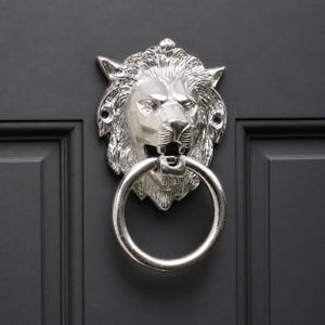 Könnte beinhalten: Ein polierter, silberfarbener Türklopfer aus Metall in Form eines Löwenkopfes, der einen kreisförmigen Ring im Maul hält. Der Löwe weist detaillierte Mähnenstrukturen und Gesichtszüge auf, mit sichtbaren Schraublöchern an den Seiten zur Montage.