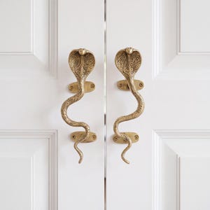 May include: A pair of brass door handles shaped like cobras with intricate engraved patterns on the hoods and bodies. Each handle is secured to a white door with two oval mounting plates, each featuring two visible screws for installation.