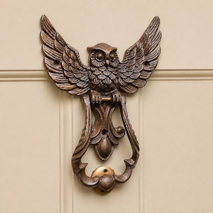 May include: A bronze-colored metal door knocker shaped like an owl with its wings spread wide. The owl features detailed feather textures and large eyes. A hinged, ornate, U-shaped ring hangs from the owl's talons, centered above a small circular mounting plate.