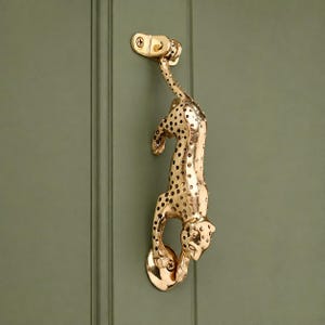 May include: A polished gold-toned metal door knocker shaped like a leaping leopard with dark spots. The leopard is attached to the door at the top by its tail and at the bottom by its front paws, featuring a hinged mechanism for knocking.