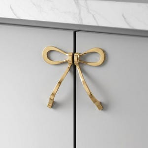 Op de afbeelding: Een paar goudkleurige metalen kastgrepen in de vorm van een strik, gemonteerd op twee aangrenzende lichtgrijze kastdeuren. Elke greep vormt de helft van de strik, met een glad, reflecterend oppervlak en gebogen, langwerpige uiteinden naar beneden.