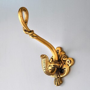 Könnte beinhalten: Ein goldfarbener Wandhaken aus Metall mit einer dekorativen Grundplatte, die mit kunstvollen Schnörkeln und einem Schraubloch versehen ist. Der Haken selbst hat ein gedrehtes Design, während der untere Teil wie eine Kobra geformt ist.