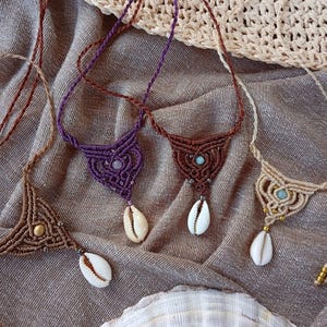 Puede incluir: Cuatro collares de macramé con intrincados diseños de cordón anudado en marrón, morado y beige. Cada collar incluye una cuenta de piedra central, pequeños detalles metálicos y un colgante de concha de cauri blanca que cuelga del nudo.