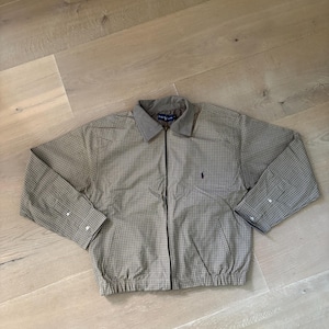 Op de afbeelding: Een beige en bruin geruite jas met ritssluiting, een effen lichtbruine kraag, lange mouwen met knoopmanchetten, een elastische zoom en een klein paars geborduurd logo op de linkerborst. Het binnenlabel vermeldt Ralph Lauren.